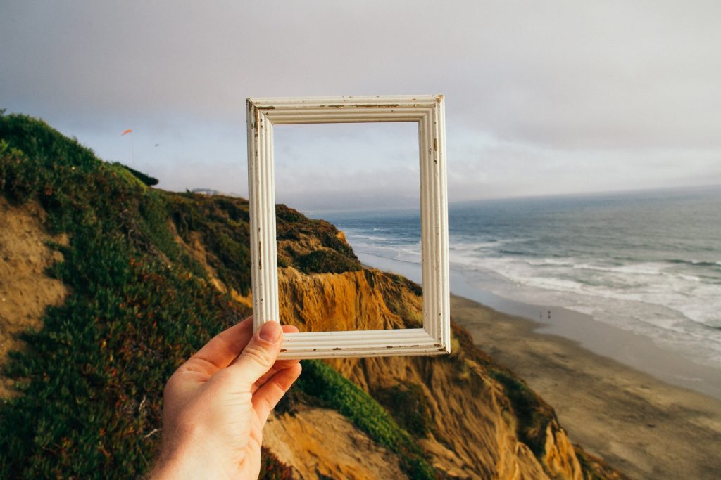 Ein Rahmen voller Möglichkeiten – diese Hand hält nicht nur einen leeren Bilderrahmen gegen die malerische Kulisse von Küste und Meer, sondern symbolisiert auch den Beginn einer Reise zu persönlicher Entwicklung und Erneuerung. Erweitern Sie Ihren eigenen Horizont durch Coaching-Resien.